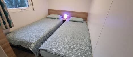 A small, single-room accommodation with two single beds, a nightstand with a lamp, and a window with a view of trees.
