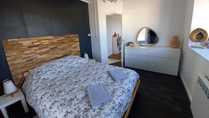 Een slaapkamer met een houten hoofdeinde, een bed met bloemenbeddengoed, een nachtkastje met een lamp, een dressoir, een spiegel en een hanglamp.