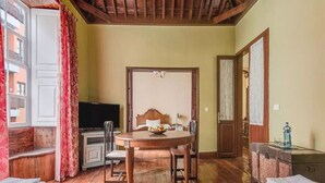 Ein Zimmer mit Holzbalkendecke, einem Esstisch mit Stühlen, einem Bett, einem Fernseher und einem Fenster mit Vorhängen.