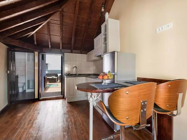 A compact kitchen with wooden cabinets, a stainless steel refrigerator, and a dining area with two wooden chairs.