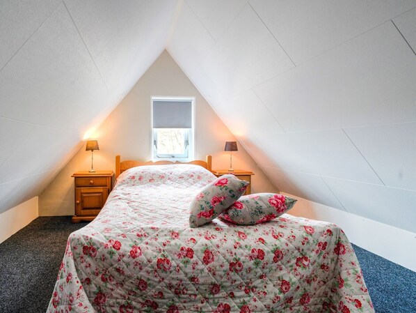 Ein Schlafzimmer mit einem Bett, zwei Nachttischen, einem Fenster und einer geneigten Decke.