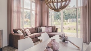 A modern living room with a sectional sofa, a dining table with vases, and large windows offering a view of the outdoors. - Modern Bungalow with Garden in Twente (Rijssen)