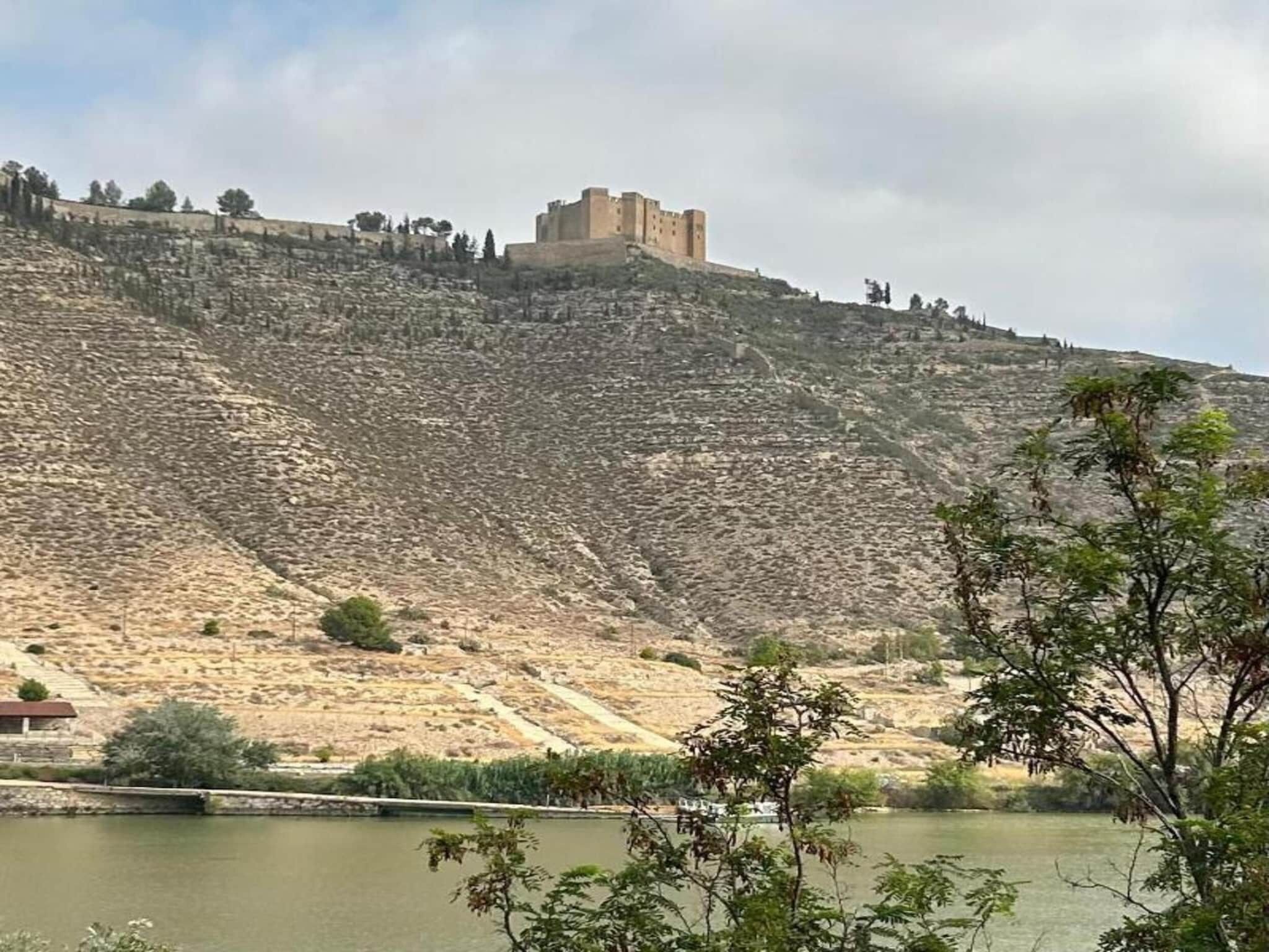 A hill with a castle-like structure on top, a body of water in the foreground, and sparse vegetation.