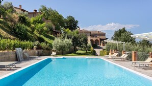 Une piscine d’un bleu éclatant, dotée de chaises longues et d’une pergola, entourée de verdure et bordée par un bâtiment situé sur une colline.