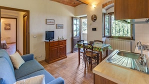 Un salon chaleureux avec un canapé bleu, un meuble en bois et un téléviseur. On y trouve également un coin repas avec une table et des chaises, ainsi qu’une cuisine équipée d’un évier et d’une fenêtre.