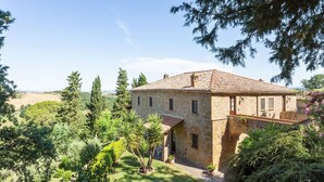 Un bâtiment en pierre, surmonté d’un toit en tuiles, entouré d’une végétation luxuriante et d’arbres.