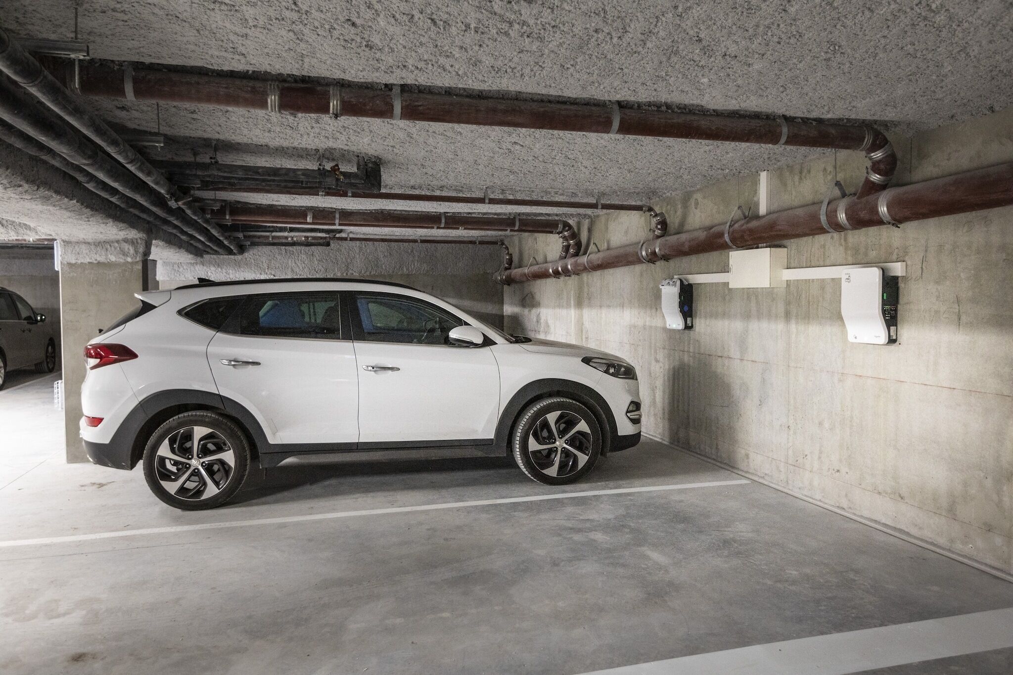 A white SUV parked in a concrete parking garage with visible pipes and a concrete ceiling.