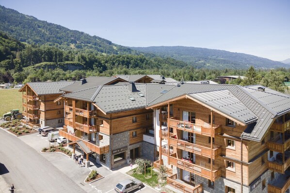 A multi-story wooden building with balconies, a parking area, and a mountainous backdrop. - Chalet in Samoëns near Grand Massif Lift (Samoëns)
