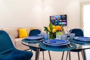 Uma sala de jantar com uma mesa redonda de vidro posta para quatro pessoas, pratos azuis e um vaso com limões e laranjas. Uma televisão de tela plana está montada na parede ao fundo.