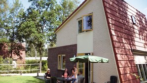 A patio area with a table and chairs, a green umbrella, and a brick building with a red roof. - Modern Villa Near Veluwe (Hoenderloo)