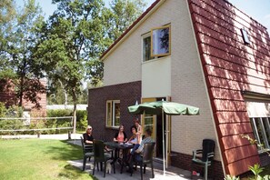 A patio area with a table and chairs, a green umbrella, and a brick building with a red roof. - Modern Villa Near Veluwe (Hoenderloo)