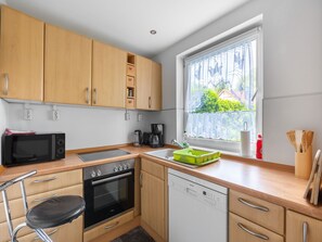 A compact kitchen with wooden cabinets, a microwave, an oven, a dishwasher, and a window with a view of greenery. - Ruhige Gartenwohnung in Dresden (Dresden)