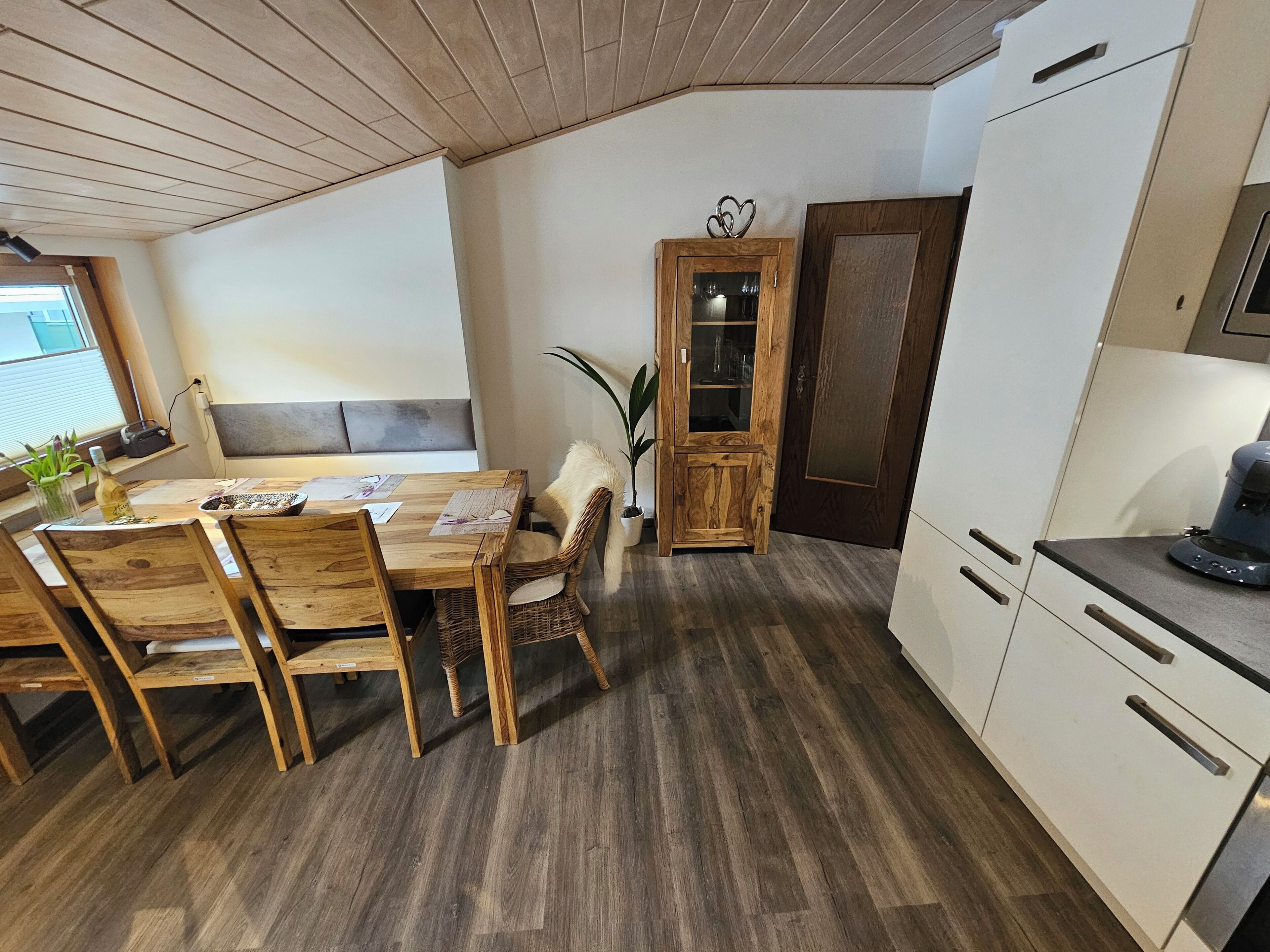 Un coin repas avec une table et des chaises en bois, un meuble avec des portes en verre et un espace cuisine avec des meubles blancs et une machine à café.
