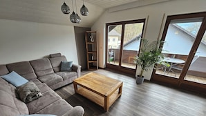 A modern living room with a sectional sofa, a wooden coffee table, and a large window offering a view of the outside. - Beautiful vacation apartment on the edge of the forest with three bedrooms. (Bad König)