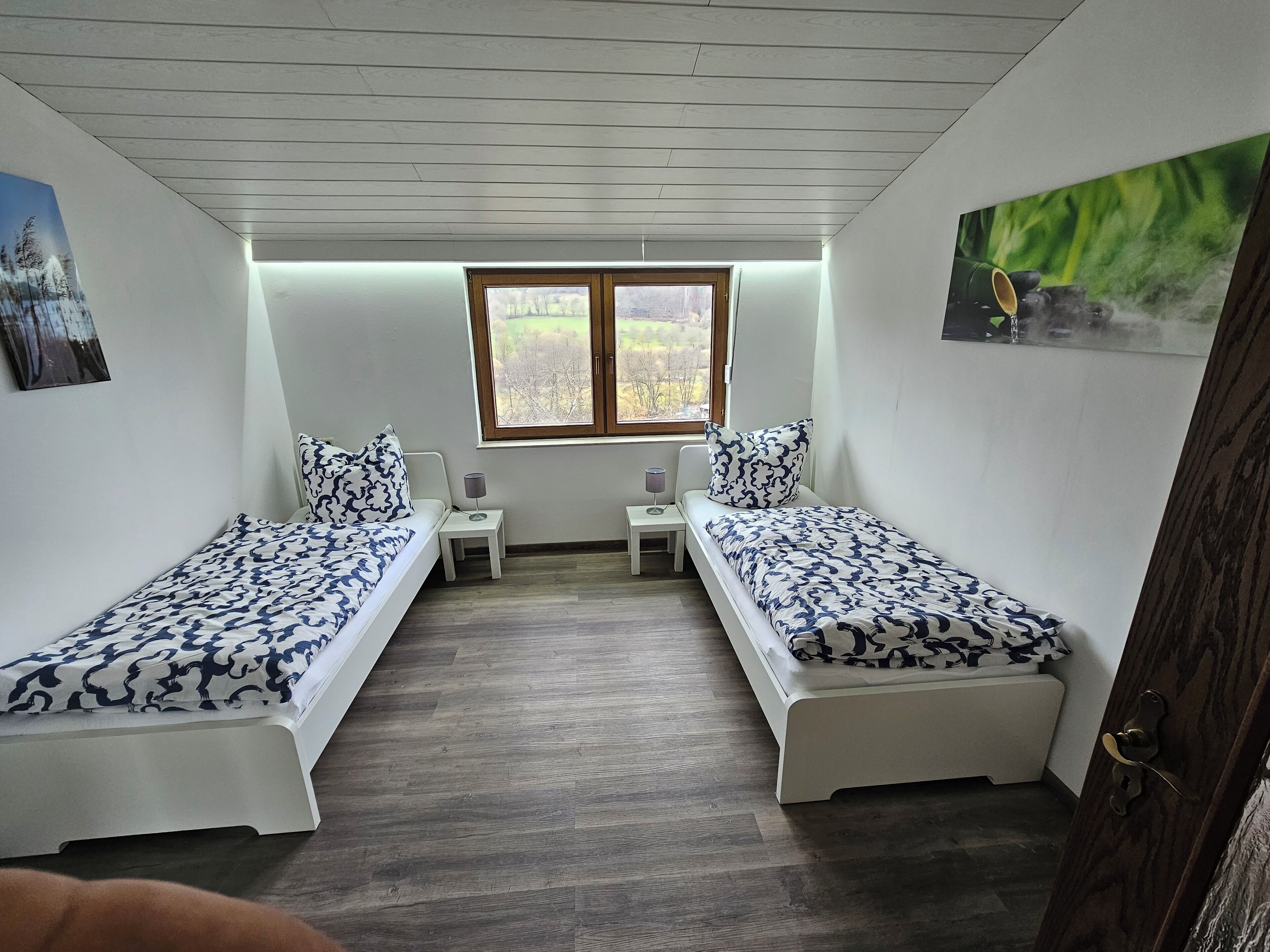 A bedroom with two single beds, a small table, a chair, and a window with a view of trees.