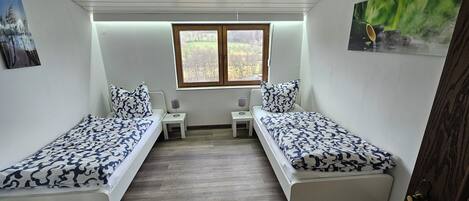 A bedroom with two single beds, a small table, a chair, and a window with a view of trees.