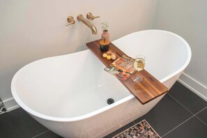 Un baño moderno con una bañera independiente, una bandeja de madera con una revista, una copa de vino y un pequeño florero con flores.