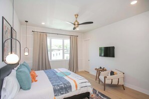 Un dormitorio con una cama, ventilador de techo, televisión, una mesita y una silla.