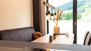 Ett modernt matområde med ett träbord, en svart vadderad bänk och utsikt över ett grönskande landskap genom stora fönster.