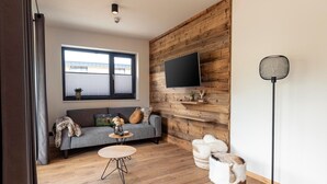 Ett modernt vardagsrum med en grå soffa, ett runt soffbord i trä, en tv som är monterad på en väggpanel i trä och en golvlampa.