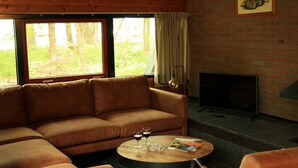 A cozy living room with a brown leather sofa, a fireplace, and a large window overlooking a forest. - Forest Hideaway with Fireplace (Herpen)