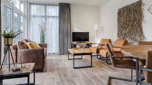 Un salon moderne comprenant un canapé, une table basse et des fauteuils, un téléviseur à écran plat et une grande fenêtre avec des rideaux.