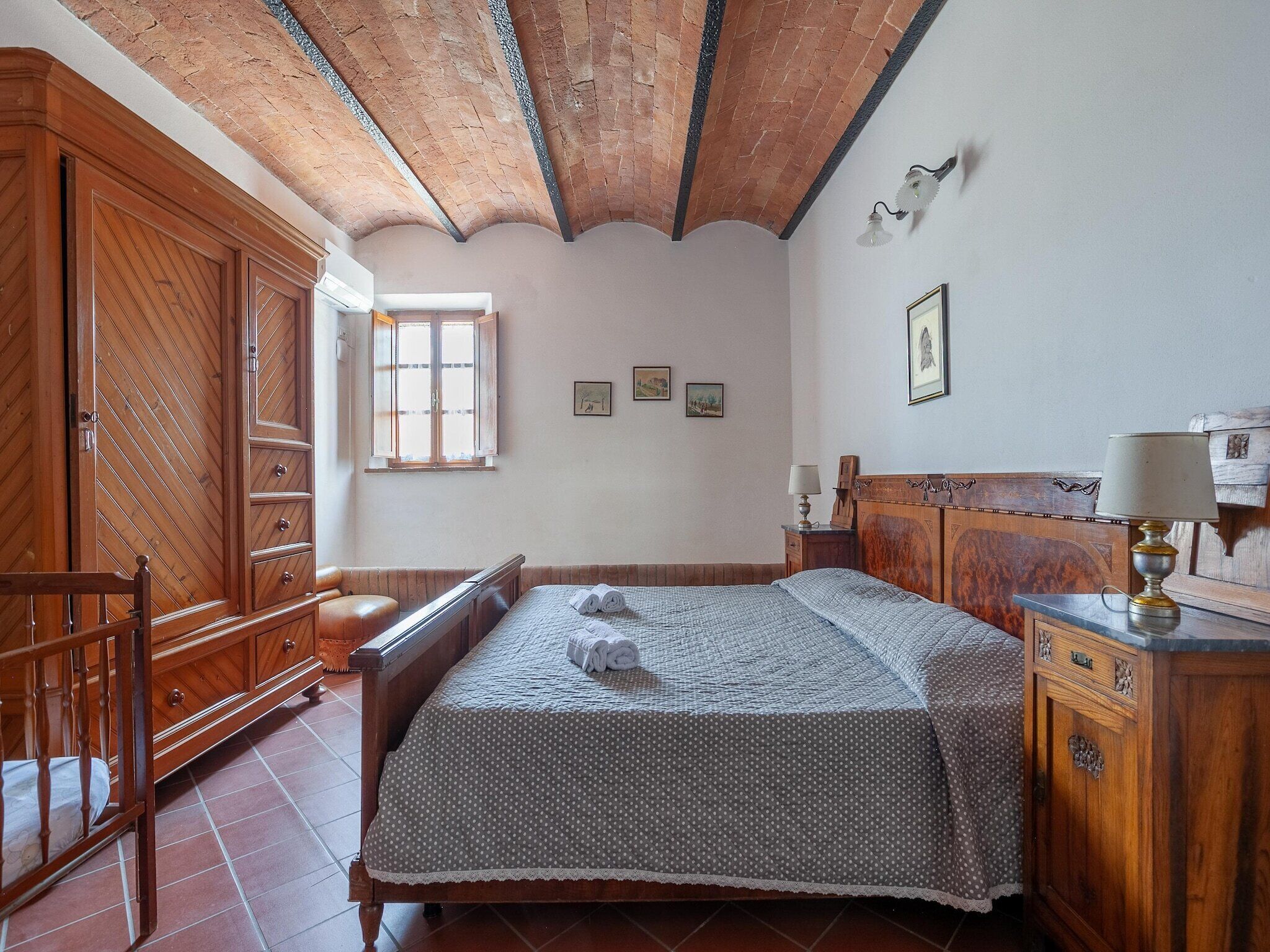 A bedroom with a wooden wardrobe, a bed with a polka dot bedspread, a nightstand with a lamp, and a window with curtains.
