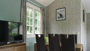 A room with a television on a wooden cabinet, a window with curtains, a chair, and a picture on the wall. - Chalet in Stegeren near Hiking Trails (Stegeren)