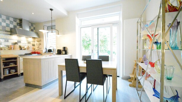 Een moderne keuken met een centraal eiland, twee zwarte stoelen, een eettafel en een glazen vitrine met kleurrijke vazen.
