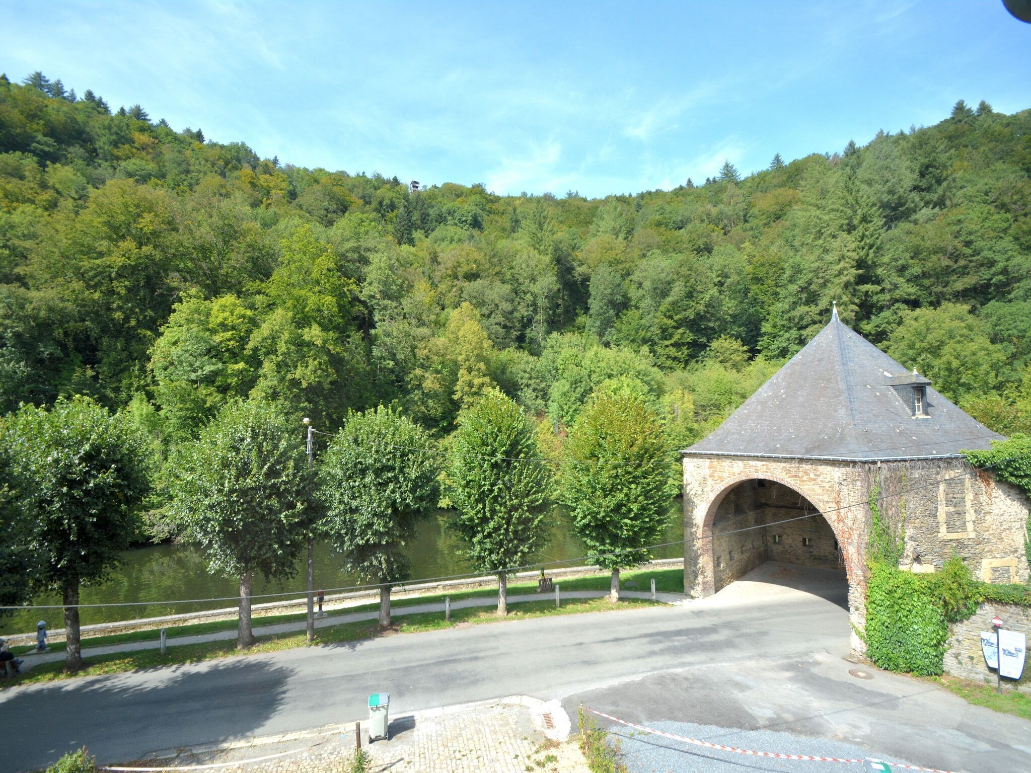 A stone bridge with a pointed roof, surrounded by lush green forest and a body of water.