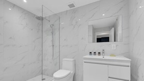 Un baño moderno con ducha de vidrio, inodoro blanco y tocador con espejo y artículos de aseo.