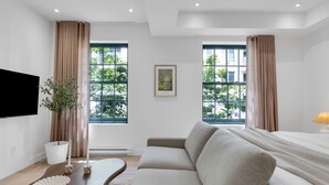 Una habitación de hotel moderna con sofá, televisor de pantalla plana, un cuadro y ventanas amplias con cortinas.