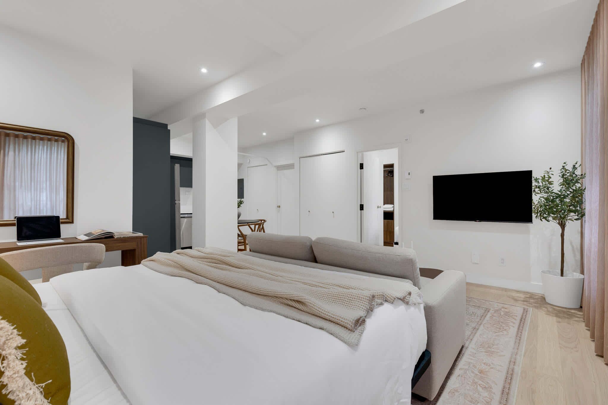 A modern hotel room with a large bed, a desk with a laptop, a flat-screen TV, and a potted plant.
