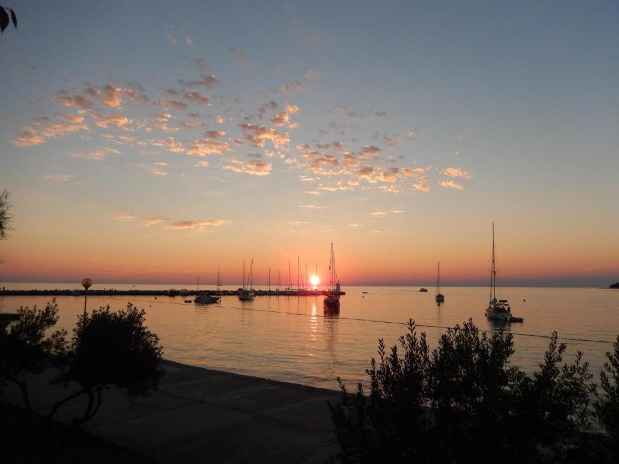 Vista do pôr do sol sobre um mar calmo, com vários barcos à vela ancorados.