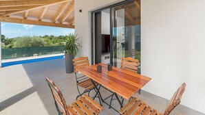 A covered outdoor dining area with a wooden table and chairs, a potted plant, and a view of a swimming pool and greenery. - Villa in Novigrad with Private Pool and Beach (Novigrad)