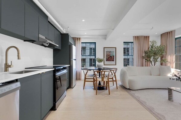 Une cuisine moderne avec des meubles gris, un plan de travail blanc et un four encastré. On y trouve un coin repas avec une table ronde et des chaises, ainsi qu’un canapé blanc dans le salon.