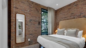 A bedroom with a brick wall, a large mirror, a bed with white bedding, and a window with curtains. - Mercer-2BDR 2BA Old Montreal- Free parking (Montréal)