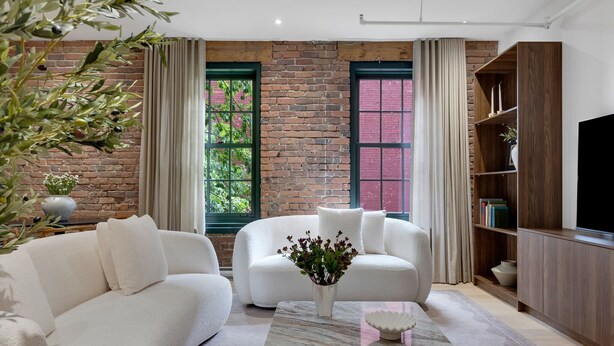 A modern living room with a brick wall, two windows with curtains, a white sofa, a coffee table with a vase, and a wooden bookshelf. - Mercer-2BDR 2BA Old Montreal- Free parking (Montréal)