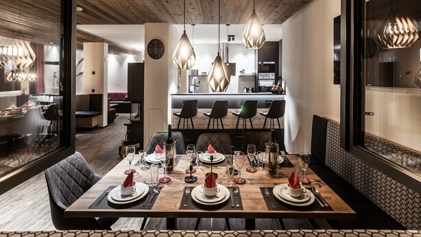 A modern dining area with a wooden table set for a meal, a kitchen in the background, and a large window with a view. - Luxury Chalet Gaschurn near Ski Lift (Gaschurn)