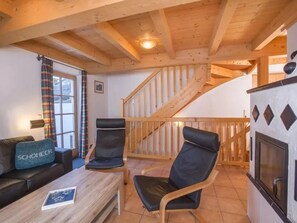 Ein gemütliches Zimmer mit Holzbalkendecke, einer Couch mit Kissen, zwei Stühlen, einem kleinen Tisch, einem Kamin und einer Treppe.