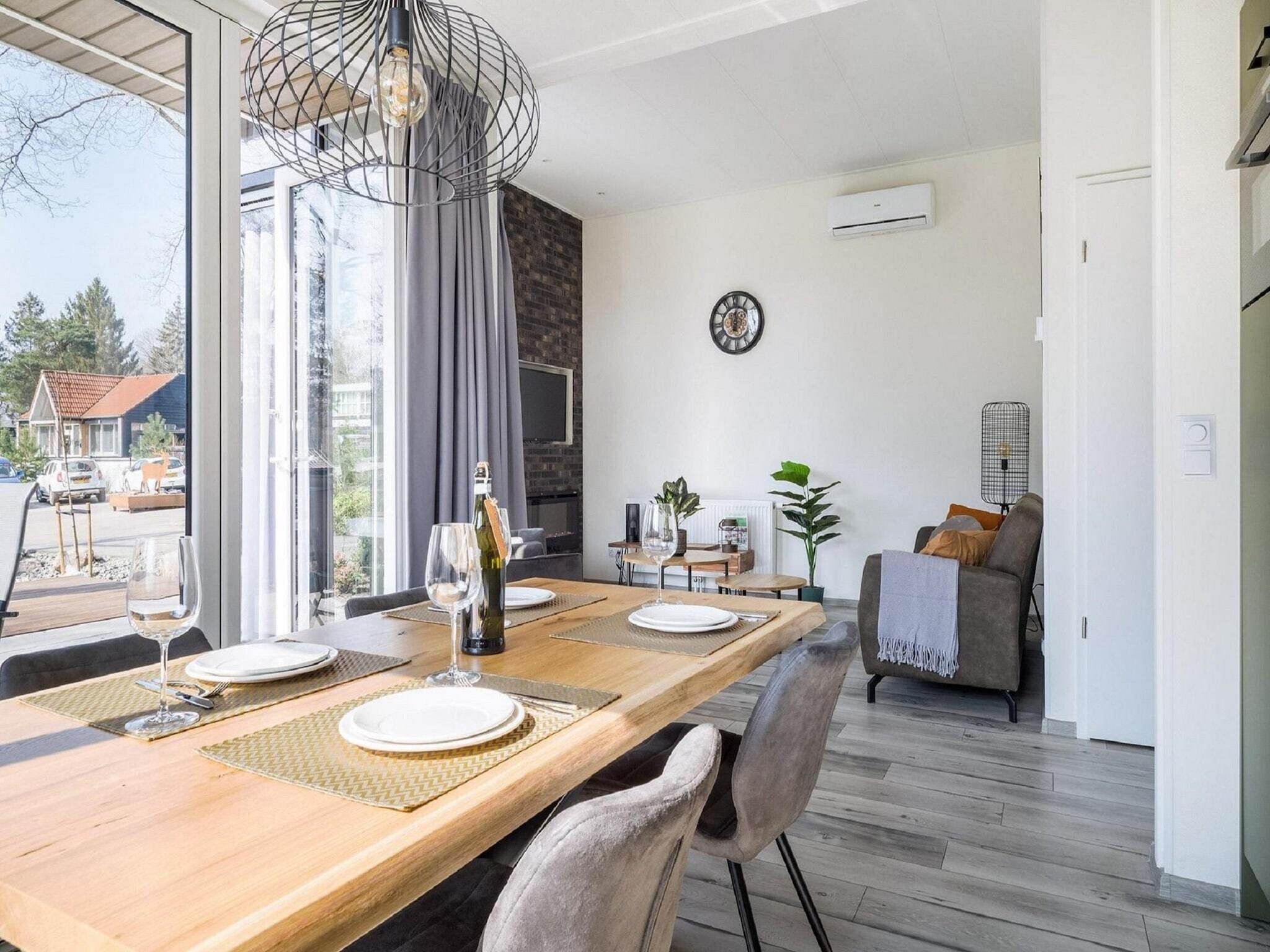 Un comedor con una mesa de madera preparada para dos personas, una botella de vino y una copa. Se ve una ventana grande con cortinas, un reloj en la pared y una planta a un lado.