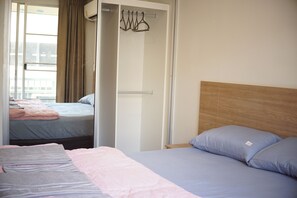 A hotel room with two beds, one with a wooden headboard and the other with a plain headboard. There is a closet with a hanging rack and a window with curtains.