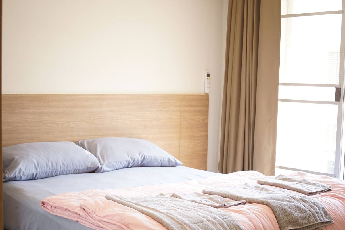A neatly made bed with a pink and blue duvet, a wooden headboard, and an air conditioning unit above it.