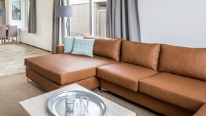 A modern living room with a brown leather sofa, a light-colored coffee table with a tray of glasses, and a dining area with chairs and a table.