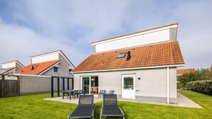 A two-story house with a tiled roof, a patio with two sun loungers, and a small outdoor seating area.