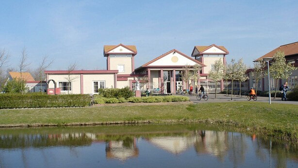 A building with a symmetrical facade, a central entrance with columns, and a reflection in a water body in front.
