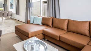 En moderne stue med en brun lædersofa, et lyst sofabord med et fad glas, og et spiseområde i baggrunden.