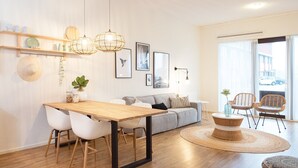 Un salon moderne avec une table à manger en bois, des chaises blanches, un canapé gris, des fauteuils en osier et une grande fenêtre donnant sur la rue.