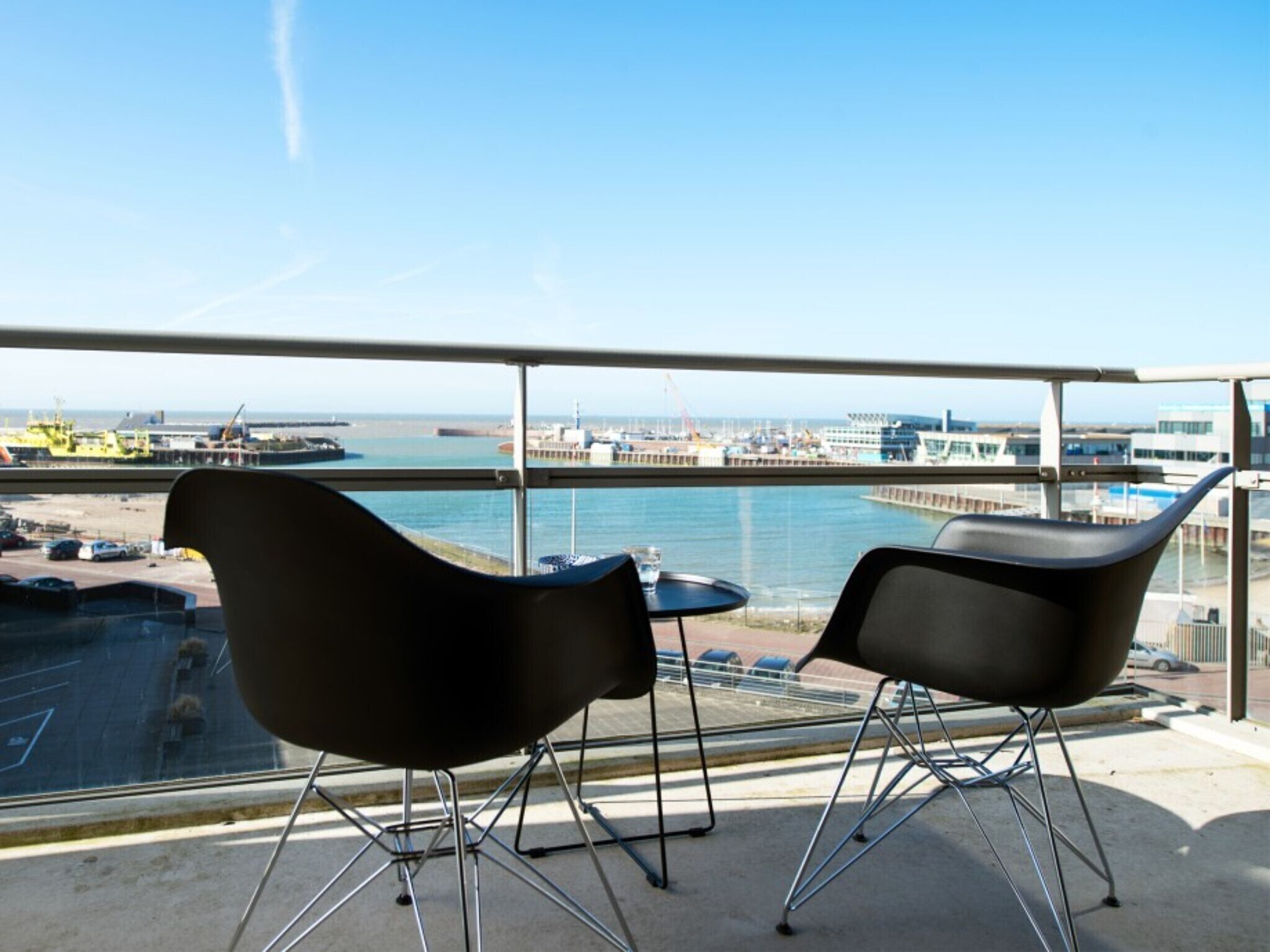 Een balkon met twee zwarte stoelen en een kleine tafel, met uitzicht op een jachthaven met boten en gebouwen.