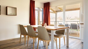 A dining area with a wooden table and chairs, large windows with red curtains, and a view of the outdoors. - Comfortable Villa in Berck (Berck-Sur-Mer)